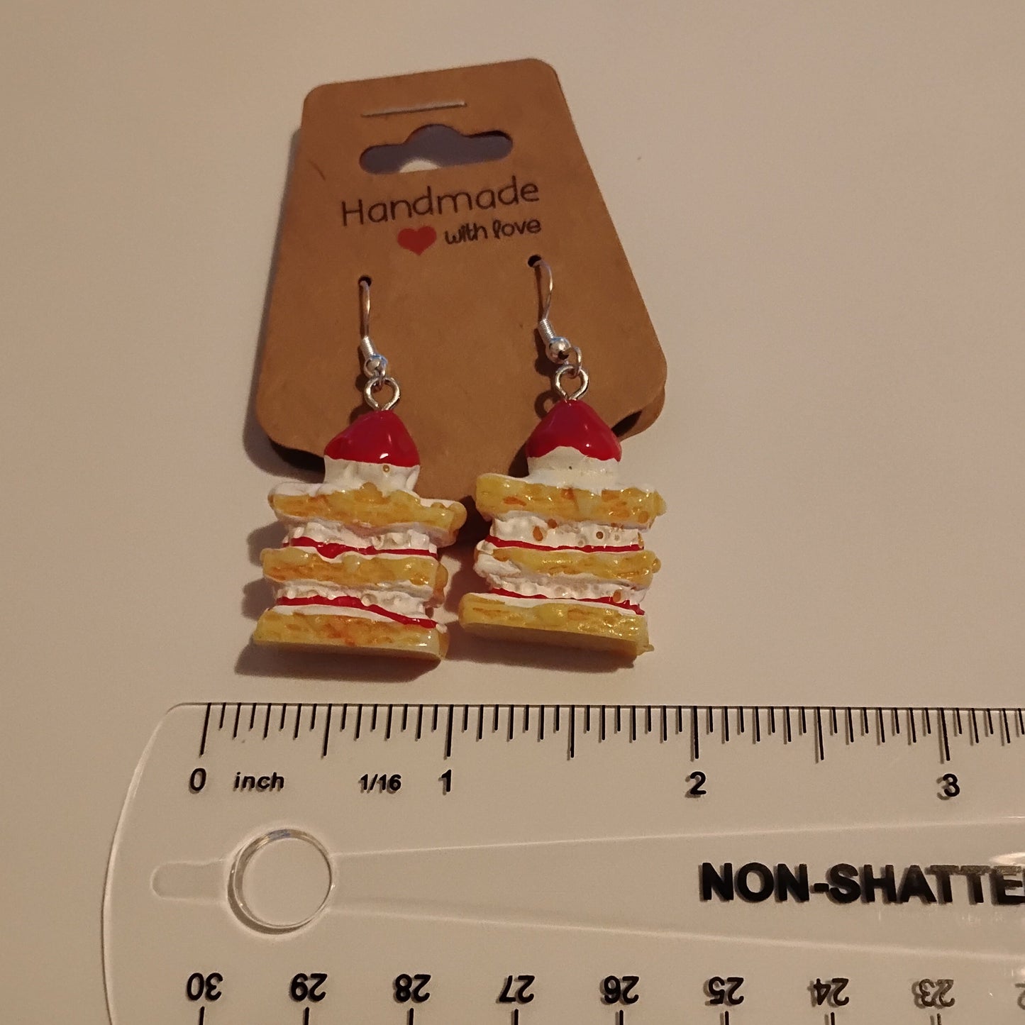 A pair of miniature sandwich-shaped dangle earrings on a brown cardboard display card, shown next to a clear plastic ruler for scale on a light beige surface.