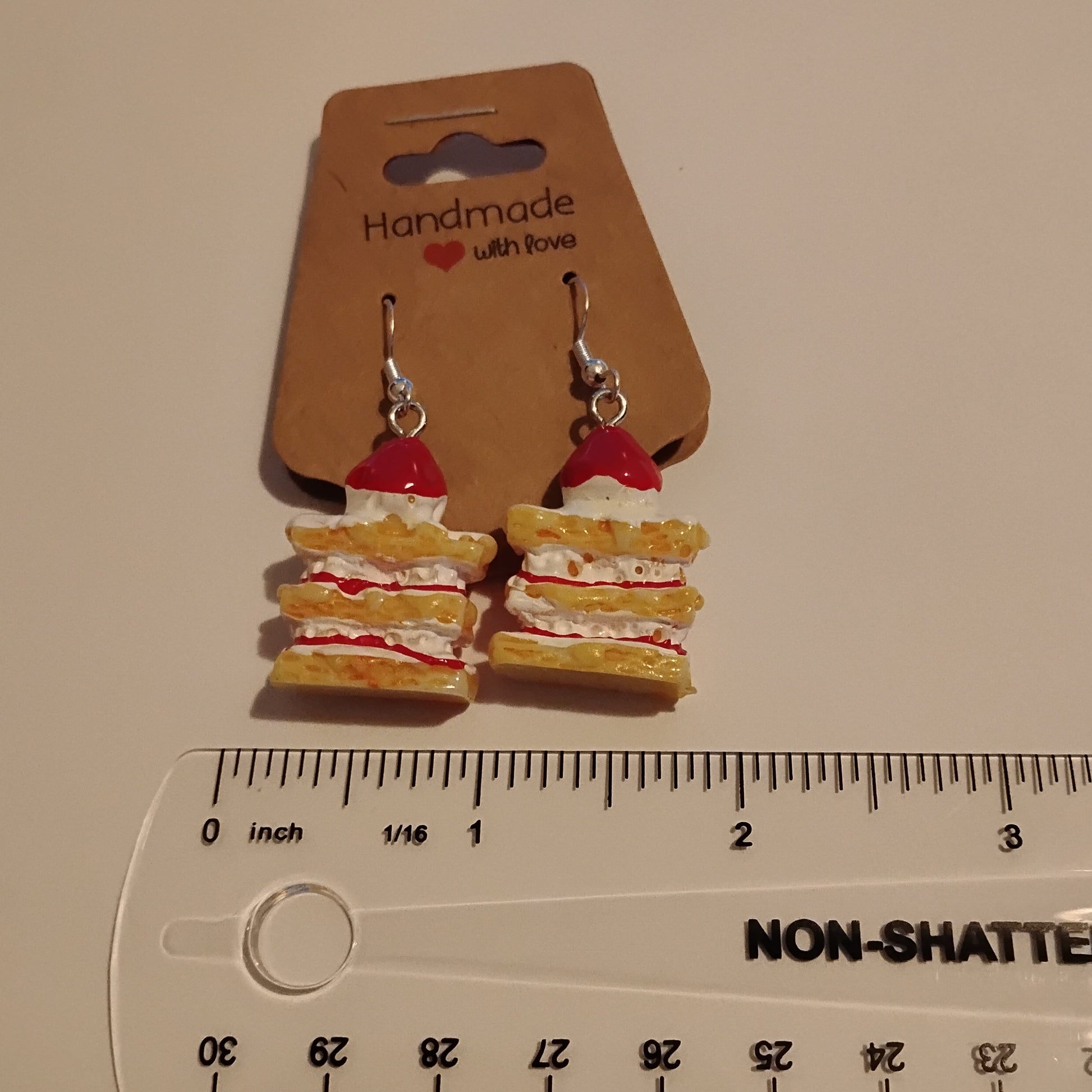 A pair of miniature sandwich-shaped dangle earrings on a brown cardboard display card, shown next to a clear plastic ruler for scale on a light beige surface.