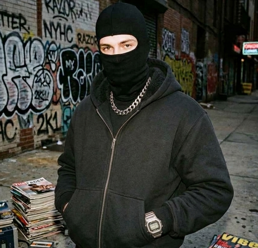 Person wearing a black hoodie and balaclava in an urban setting with graffiti.