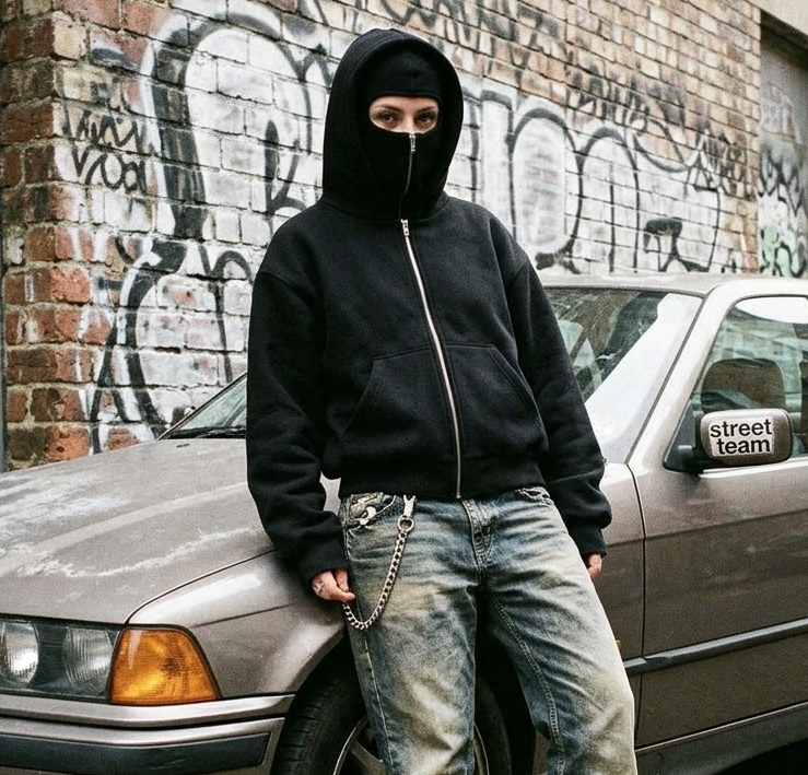 Person wearing a black hoodie and jeans standing next to a car with graffiti on the wall.